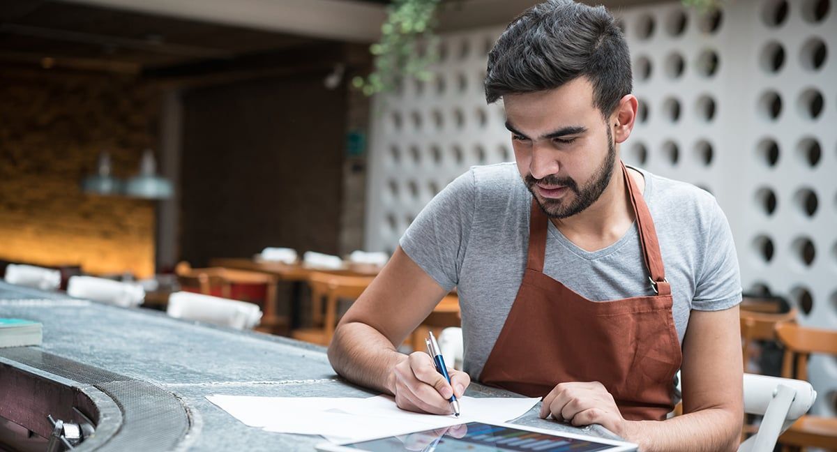 Man wearing a brown apron working at a restaurant counter, writing on paper while using a tablet.