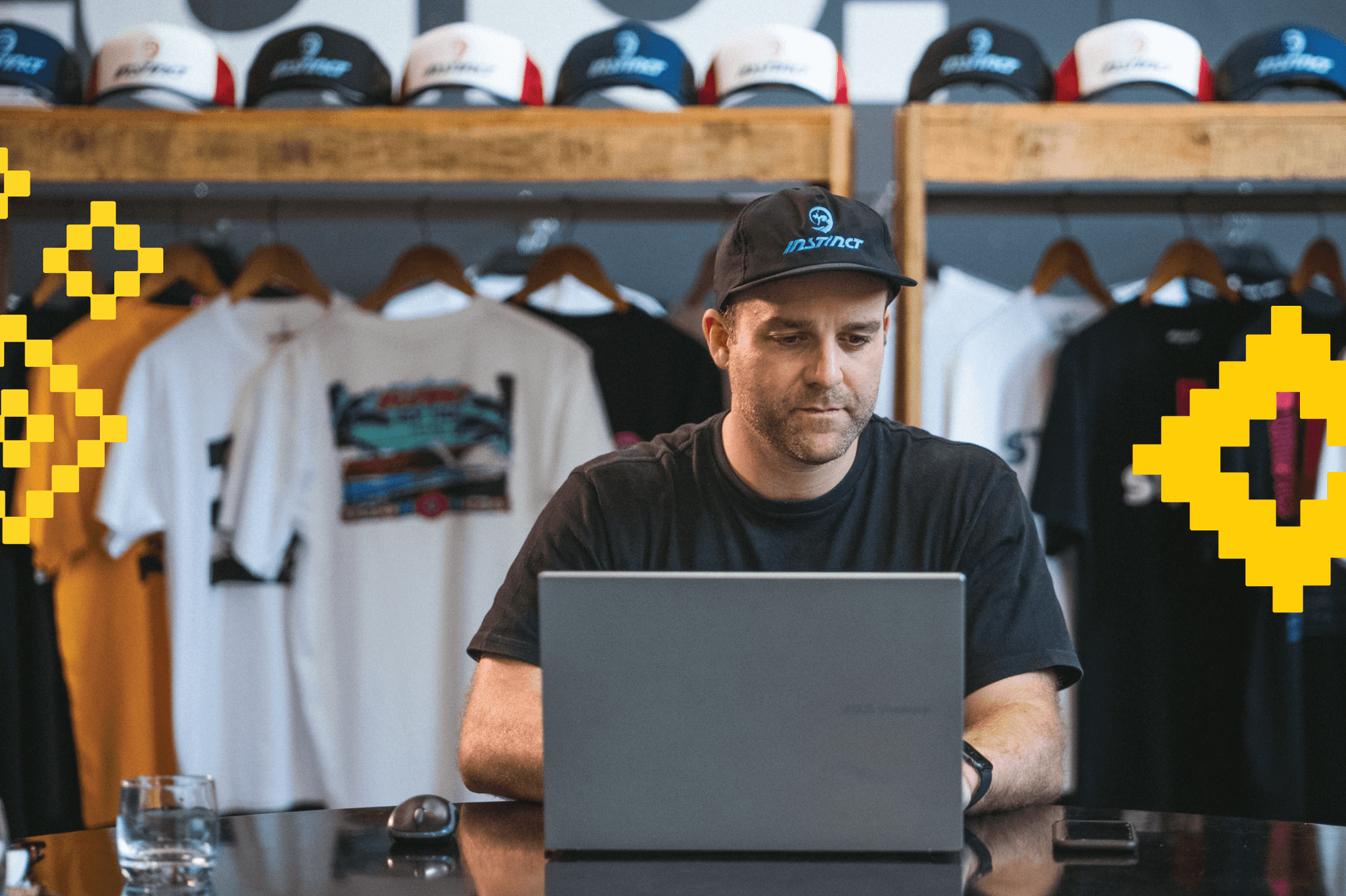 Man wearing a cap seated at a table working on a laptop, with branded hats on a shelf above and graphic t‑shirts hanging behind him